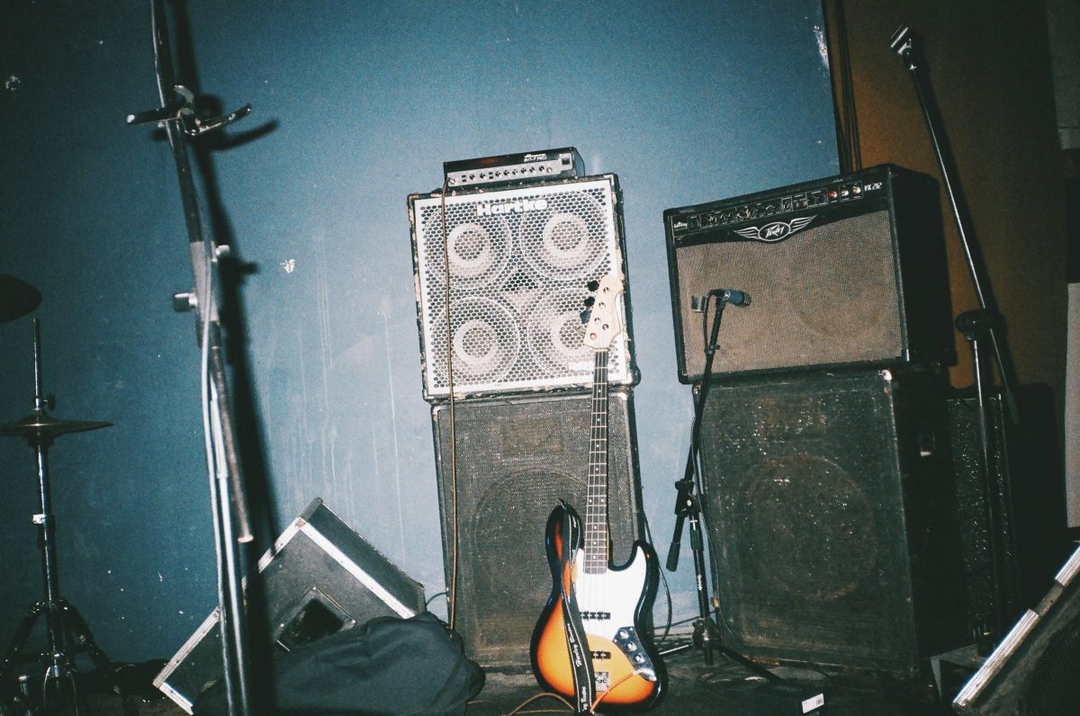 A sunburst Jazz-style bass leaning against a Hartke bass cabinet beside a Peavey combo amp on a small stage, with mic stands and a cymbal