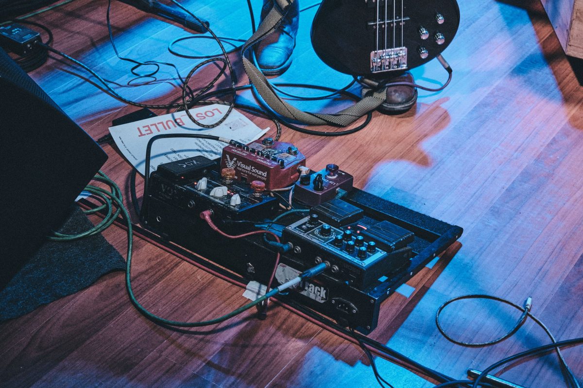 A small pedalboard on a venue floor beside a bass guitar, with a Vox pedal, Visual Sound and Boss pedals, cables and a set list