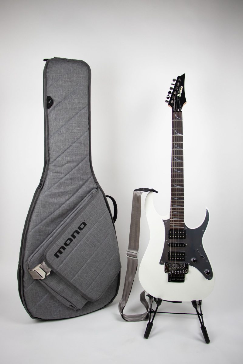 A gray padded gig bag with a large front accessory pocket, standing beside a white electric guitar on a stand