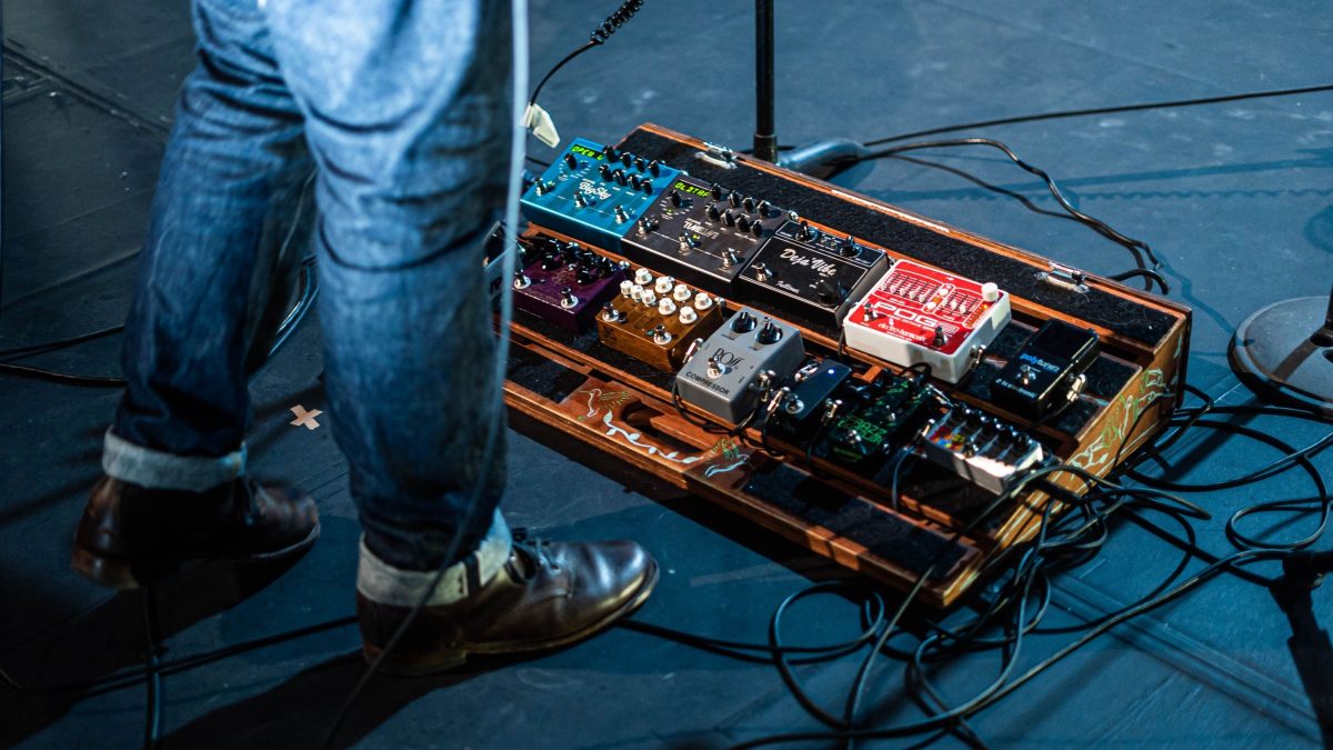 A guitarist standing beside a large wooden pedalboard with about a dozen pedals, including a compressor, on a stage floor