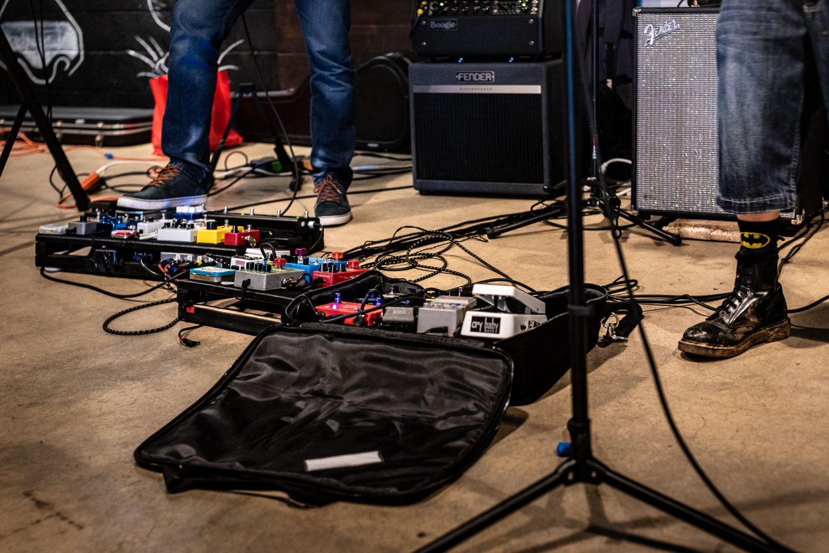Two pedalboards on a concrete venue floor, one still in its open soft case, with Fender amps and a microphone stand behind